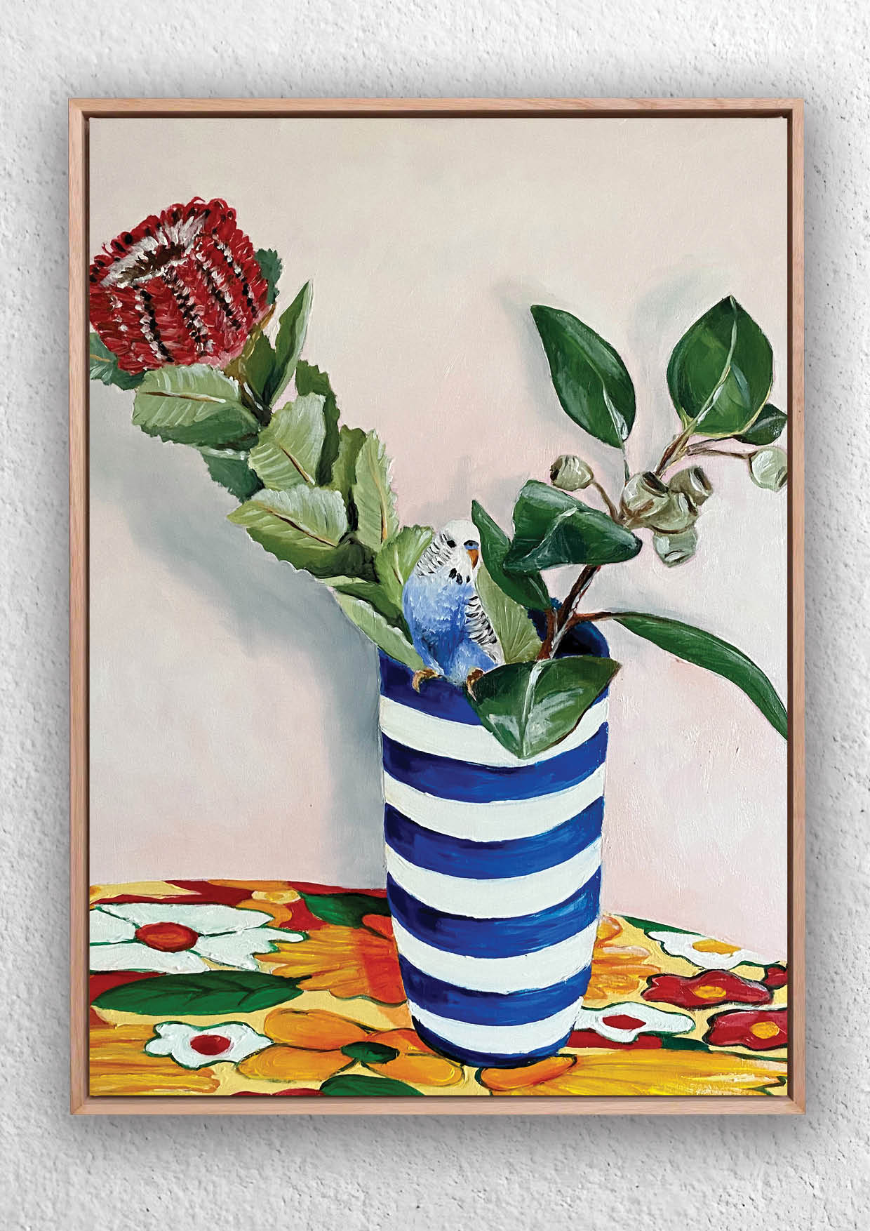 Framed artwork of a blue and white striped hamptons vase with banksia flower and gum nuts and a budgie bird on a colorful table cloth.