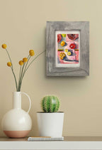 Framed original artwork on a wall of a lemon, flower and vase in reds and yellows.
